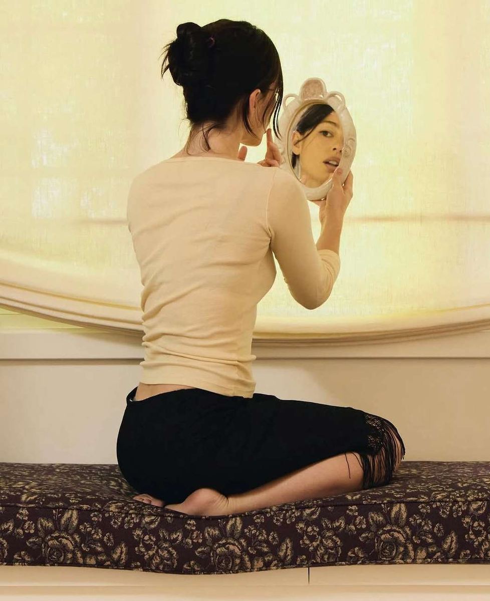 A woman seated by a window, holding a hand mirror